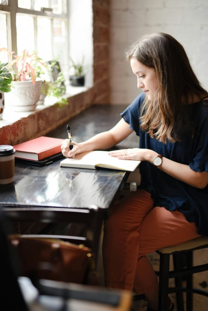 Woman at a sunlit desk writing in an open notebook with a pen